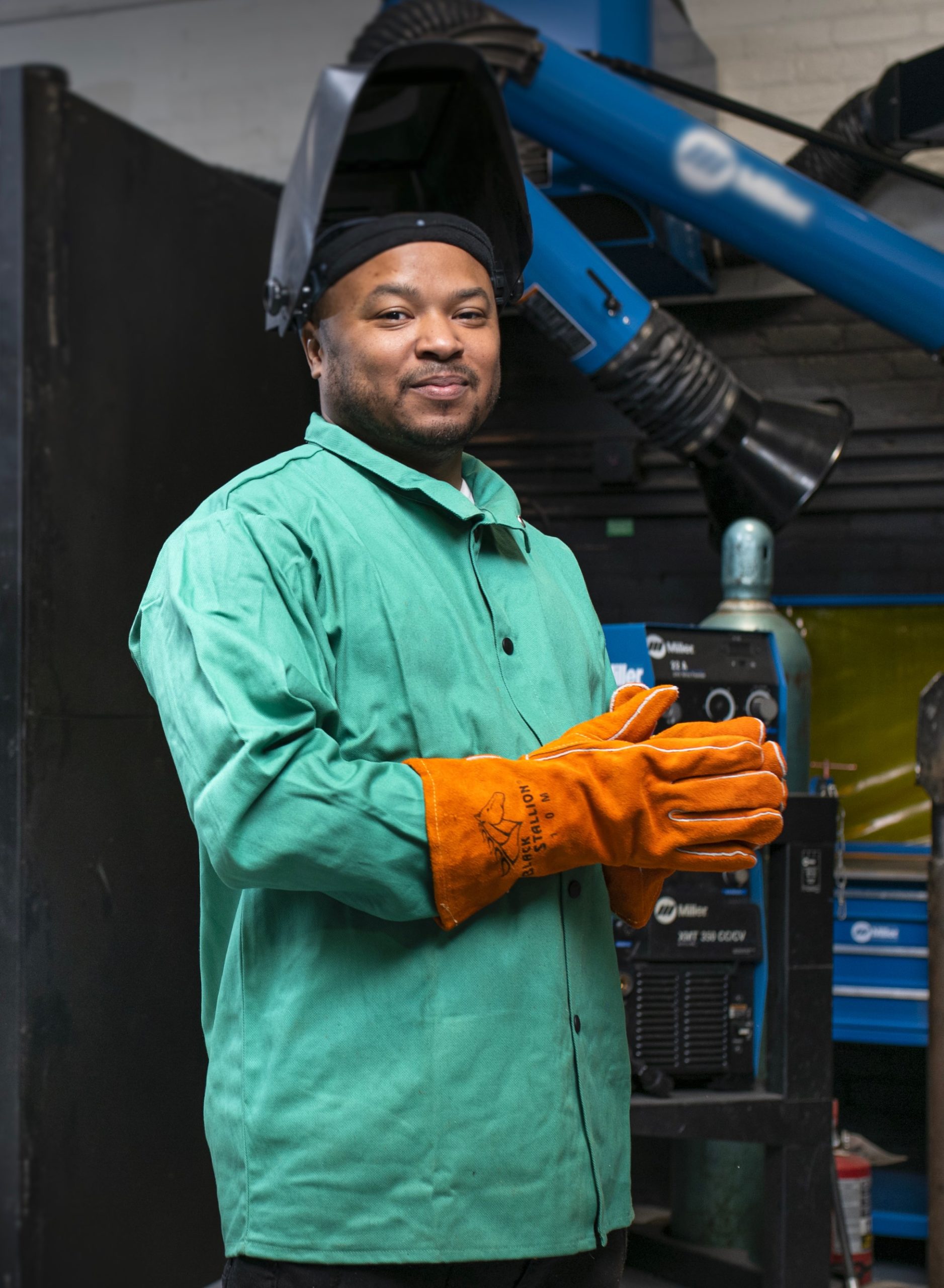 A young black man wearing a welder's mask and a green workman's suit, smiling