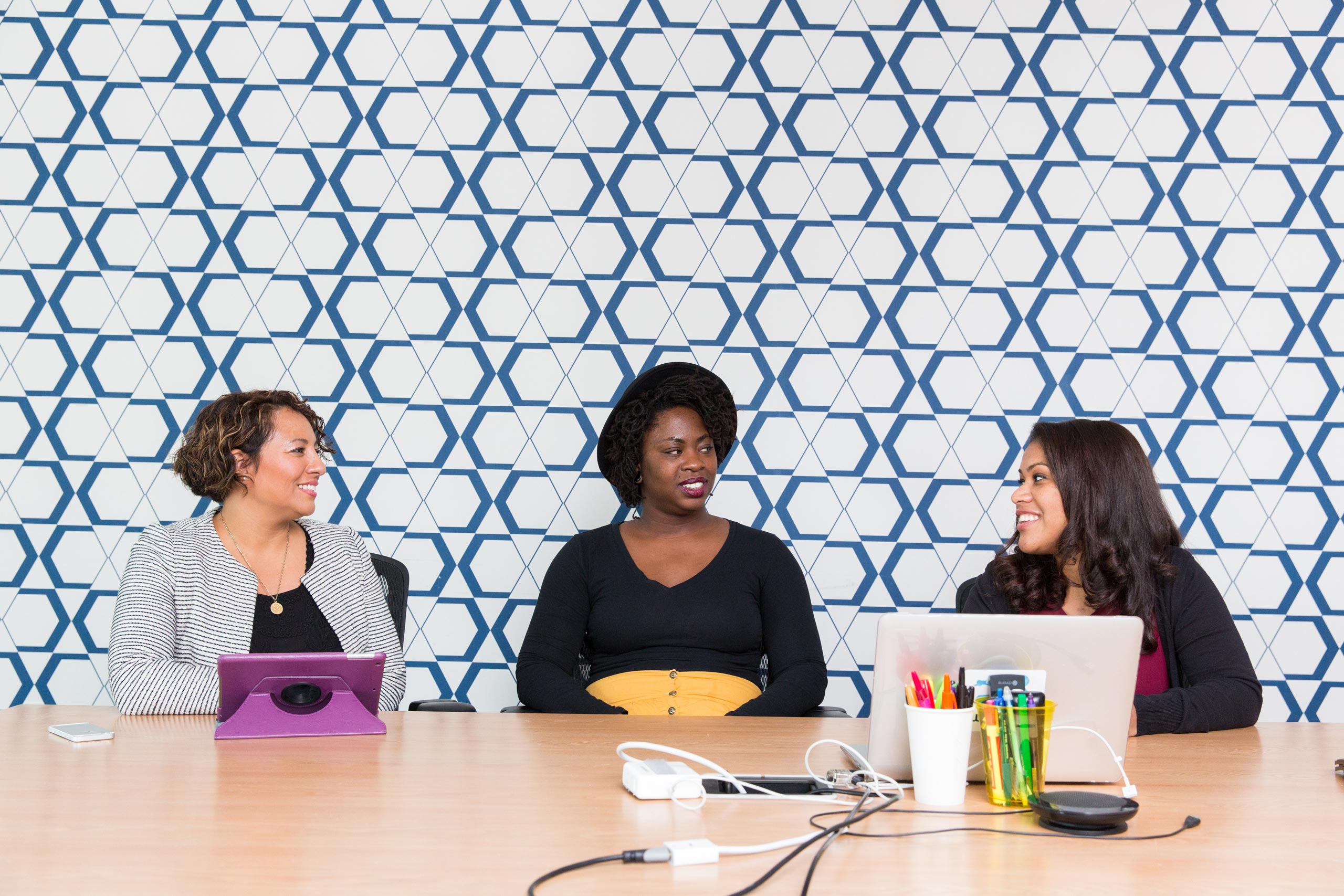 3 women consulting with each other professionally with laptops and ipads on the table in front of a blue patterned tile background