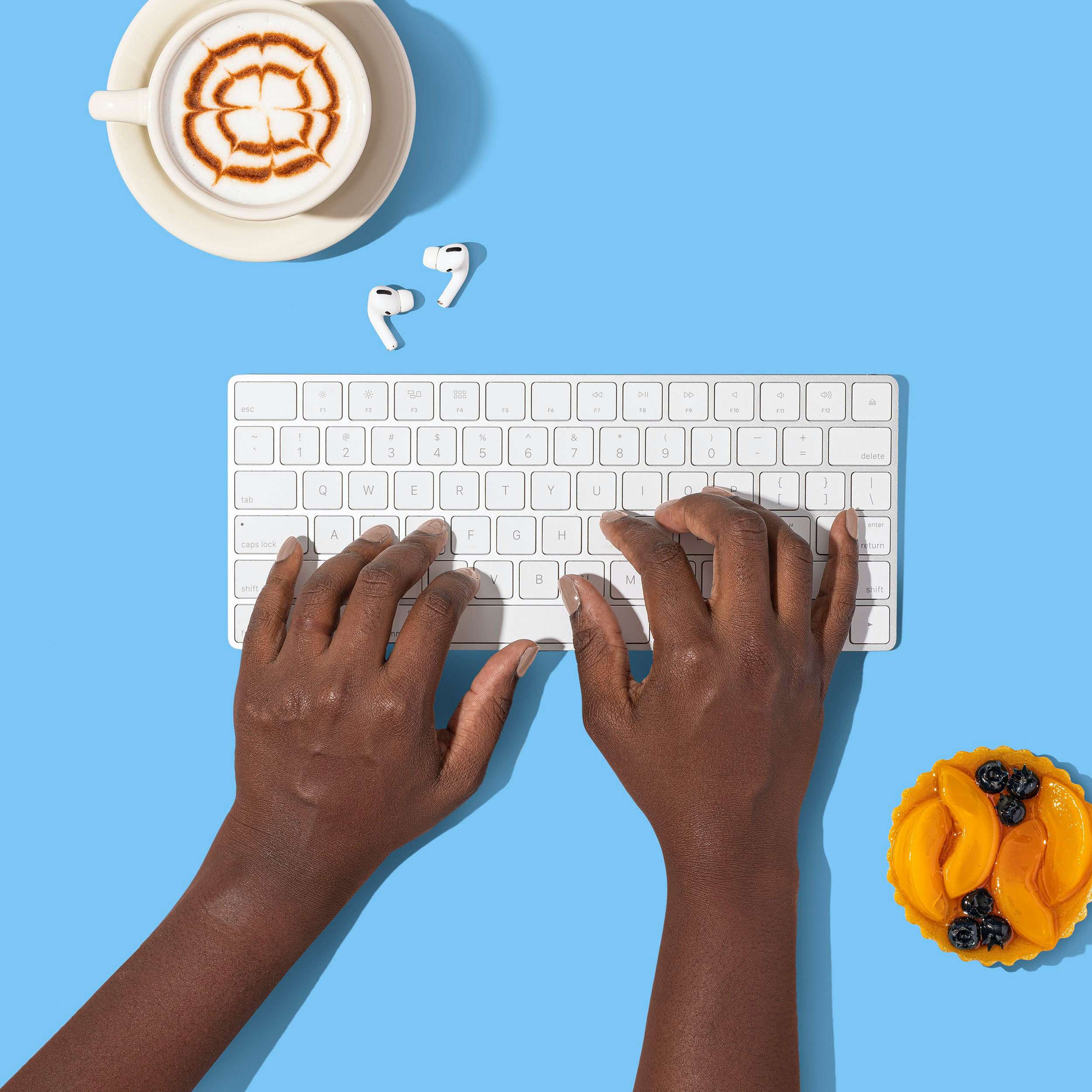 A young black woman's hands typing on a white keyboard, set on a blue background, with a cappuccino and a fruit tart nearby