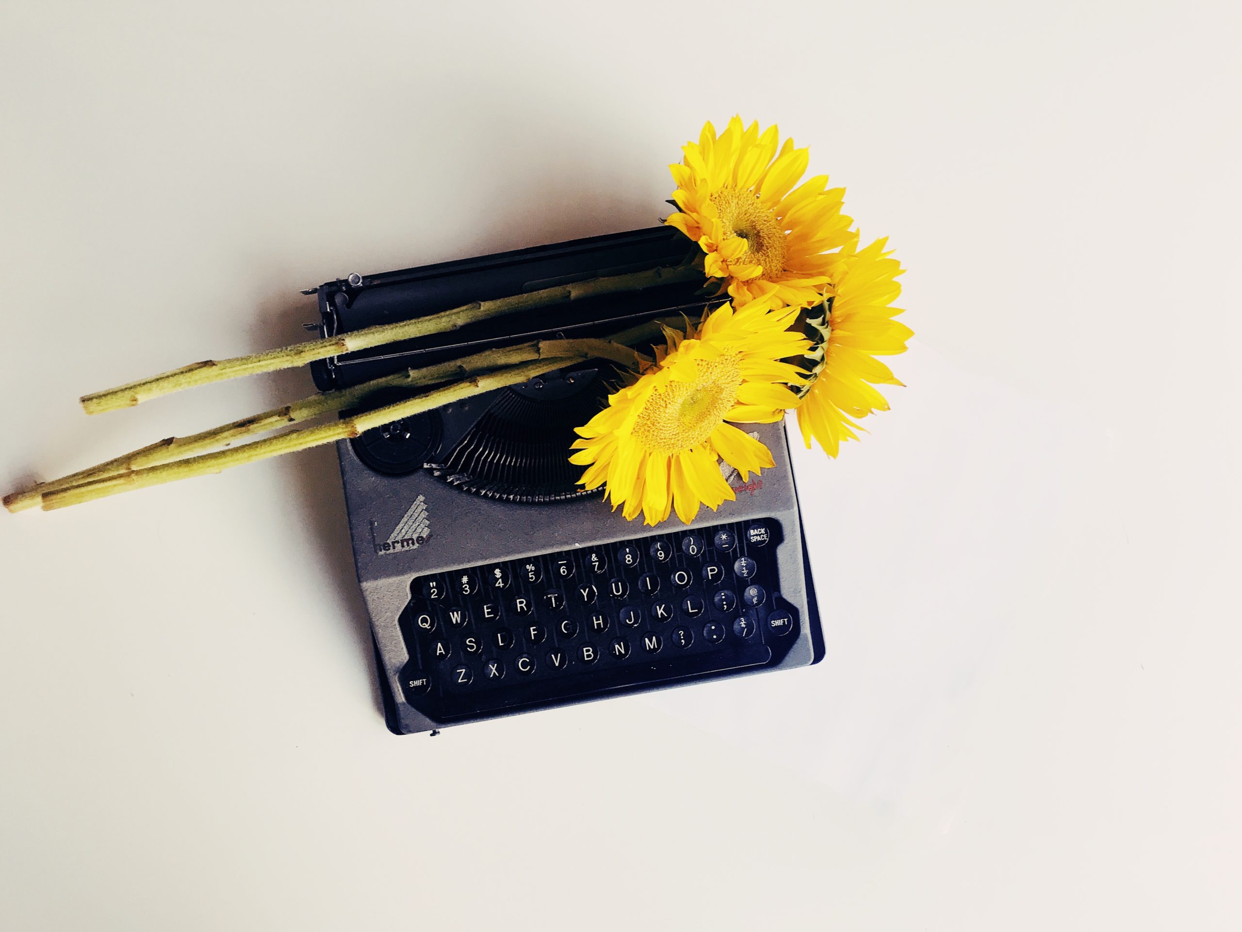 A black typewriter with yellow sunflowers set on top of it, all on a plain taupe background
